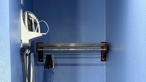 Ein Zimmer mit blauer Wand, einem Metallregal mit Kleidung, einem Metallstuhl und einem kleinen Tisch.