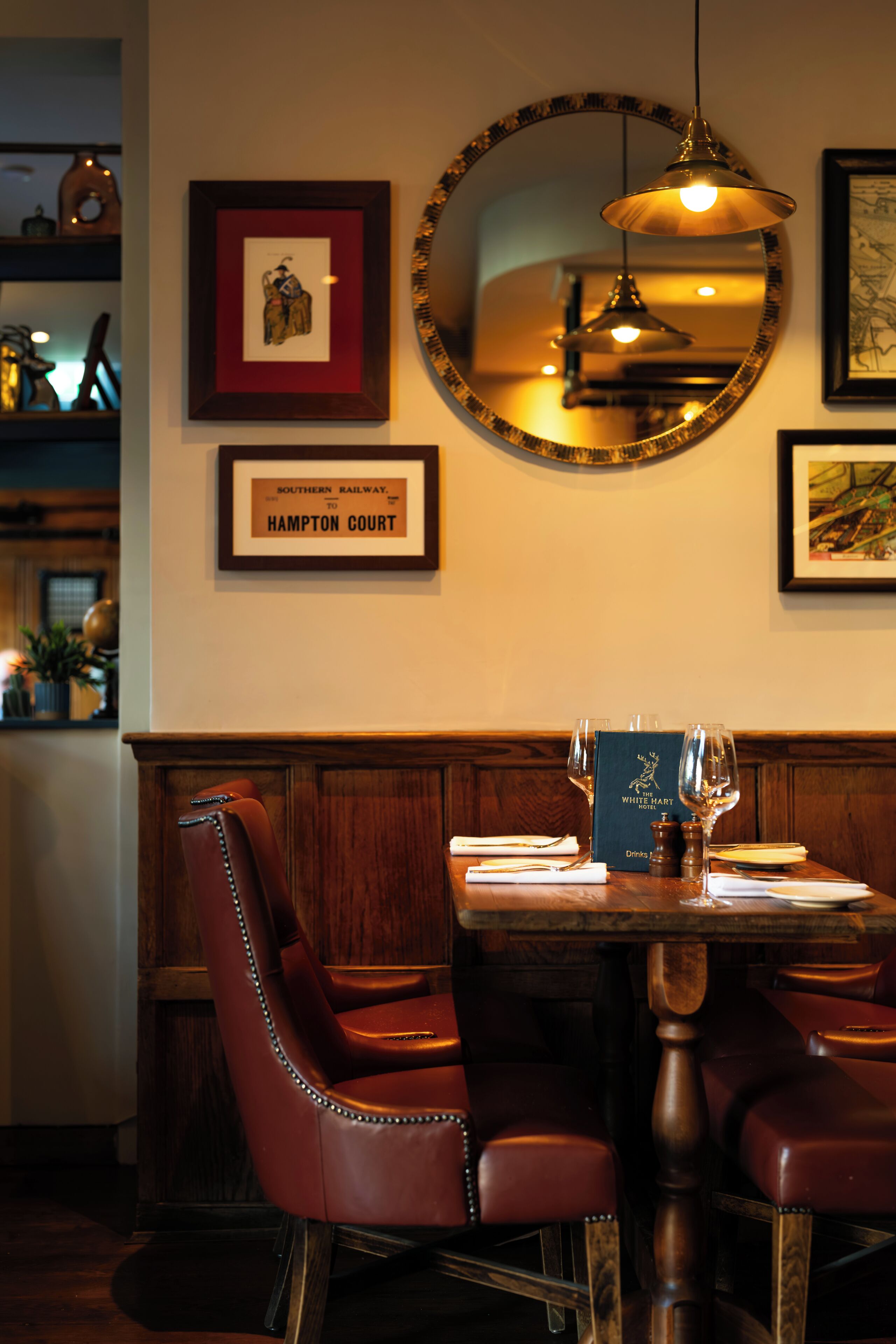 A restaurant interior with a wooden dining table, leather chairs, framed artwork, and a mirror on the wall.