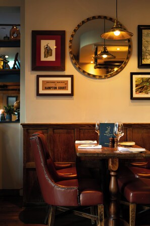 A restaurant interior with a wooden dining table, leather chairs, framed artwork, and a mirror on the wall.