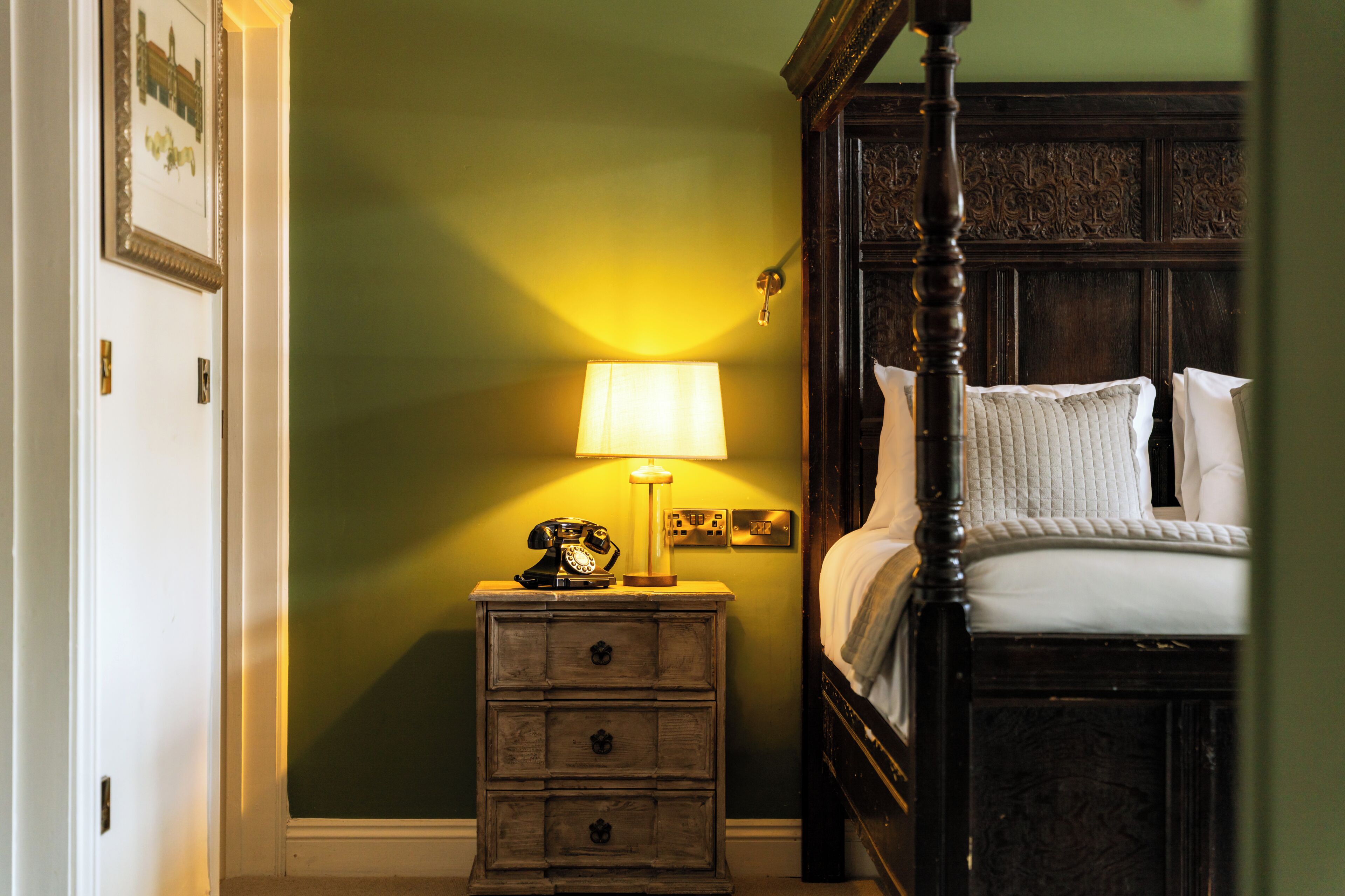 A bedroom with a wooden four-poster bed, a nightstand with a lamp, and a wall-mounted light switch.