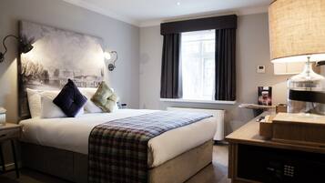 A hotel room with a large bed, a bedside table with a lamp, a window with curtains, and a thermostat on the wall.