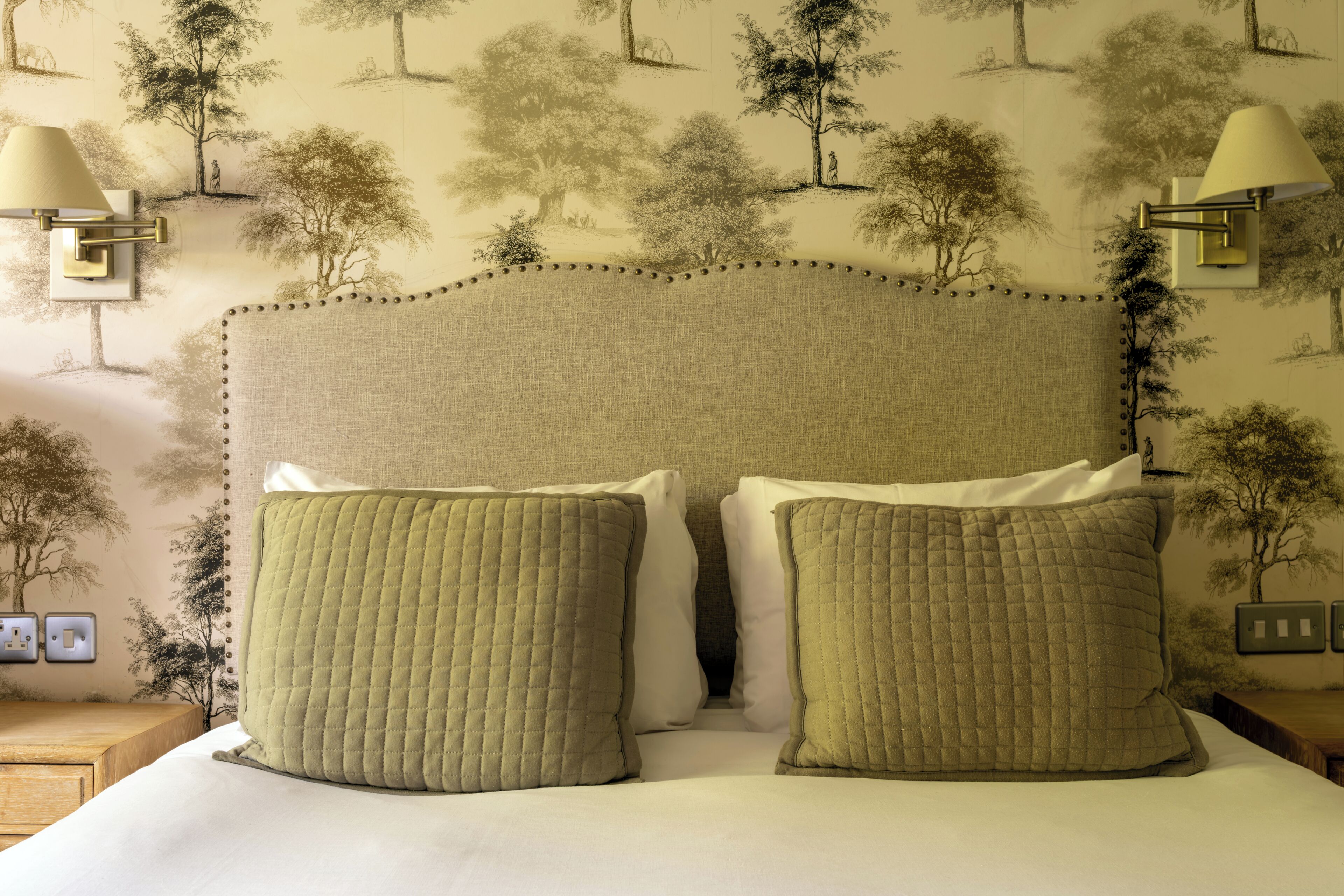 A bed with a quilted headboard and two pillows, set against a wall with a nature-themed wallpaper.