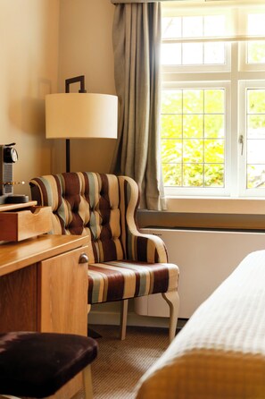 A hotel room with a bed, a desk with a lamp, a striped armchair, and a window with curtains. - The White Hart Hotel (Kingston Upon Thames)