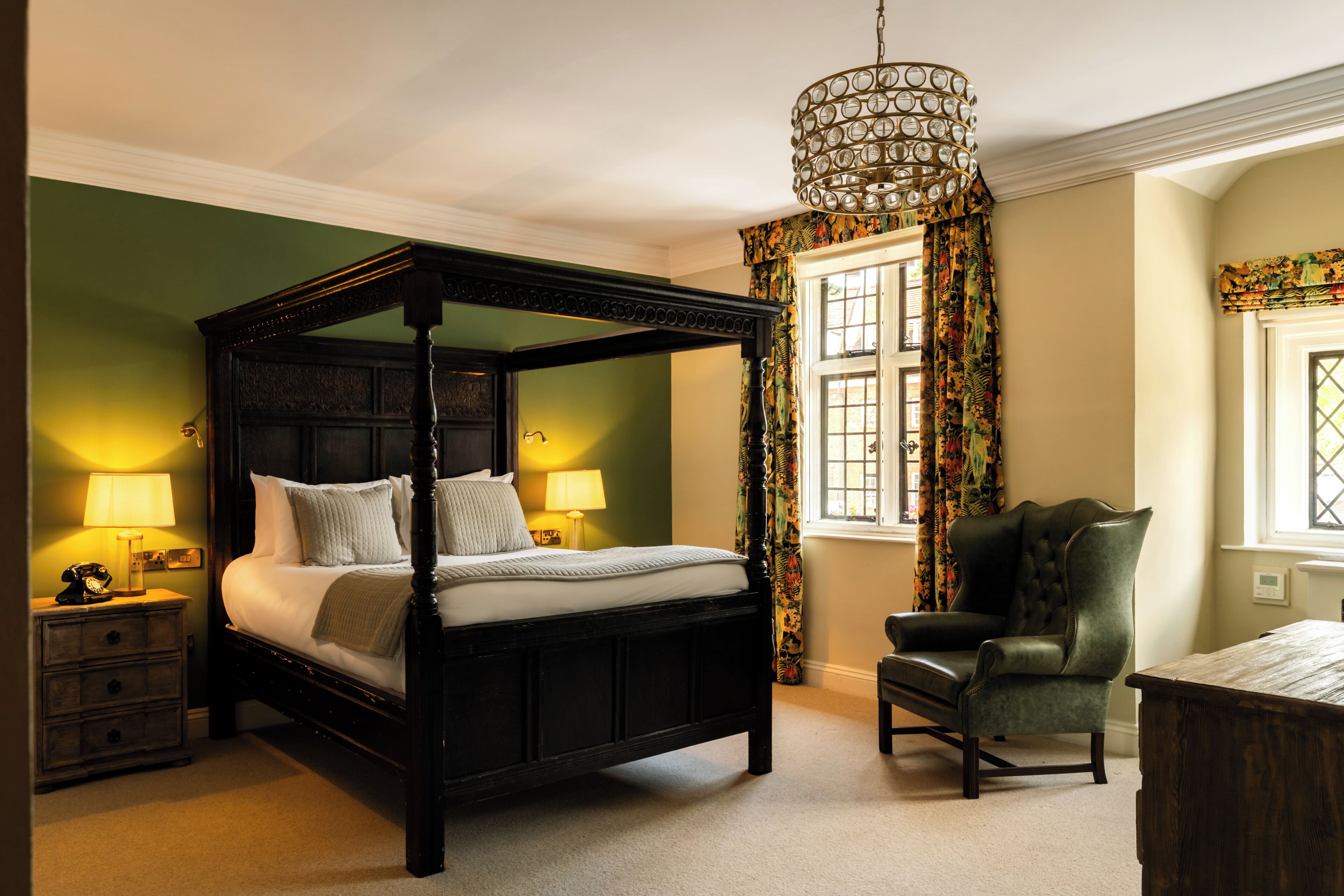 A four-poster bed with a canopy, two bedside lamps, a green chair, and a decorative chandelier.