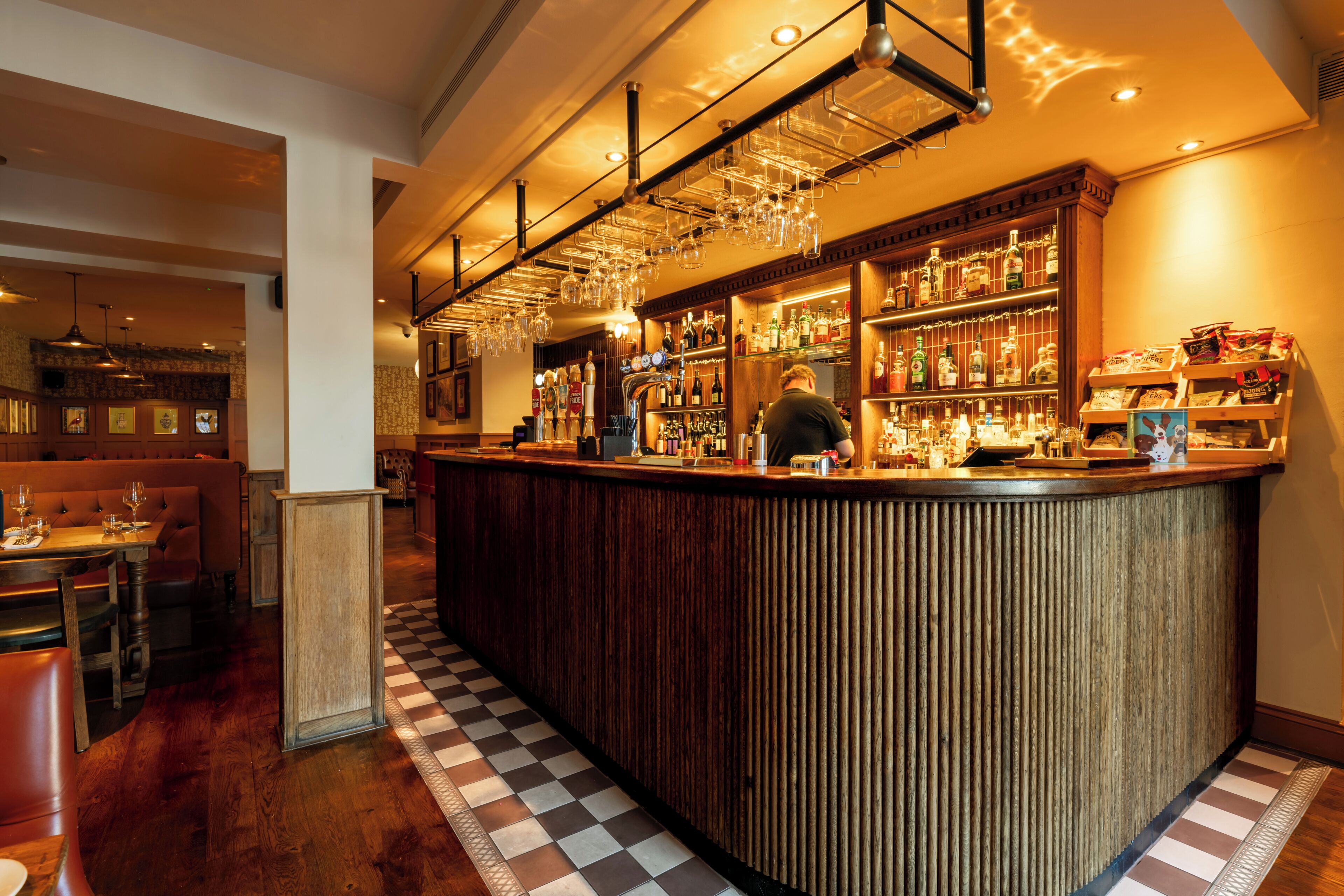 A bar area with a bartender, wooden bar counter, and shelves of bottles.