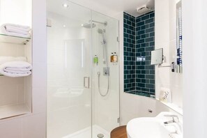 A modern bathroom with a glass-enclosed shower, white tiles, a wooden toilet, and a mirror with lights. - The White Hart Hotel (Kingston Upon Thames)