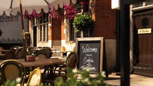 An outdoor seating area with wicker chairs and tables, a chalkboard sign welcoming guests, and a brick building with a door labeled 'To The Bar'. - The White Hart Hotel (Kingston Upon Thames)
