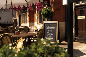 An outdoor seating area with wicker chairs and tables, a chalkboard sign welcoming guests, and a brick building with a door labeled 'To The Bar'.