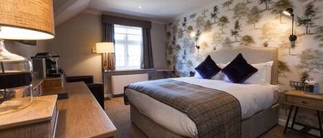 A hotel room with a large bed, a desk with a lamp, a window with curtains, and a wall with a tree pattern.