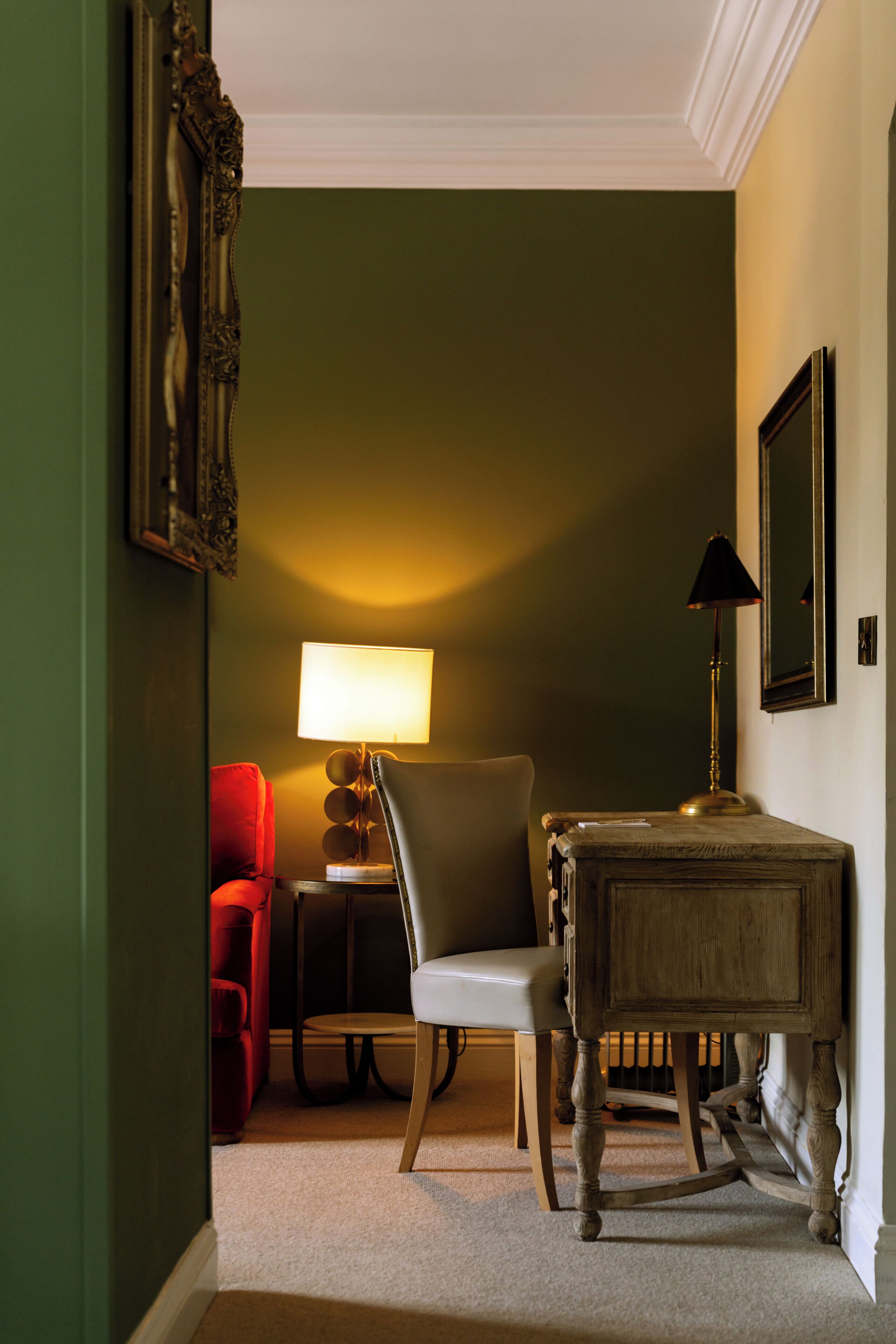 A room with a green wall, a wooden table, a chair, a red chair, a lamp, and a mirror.