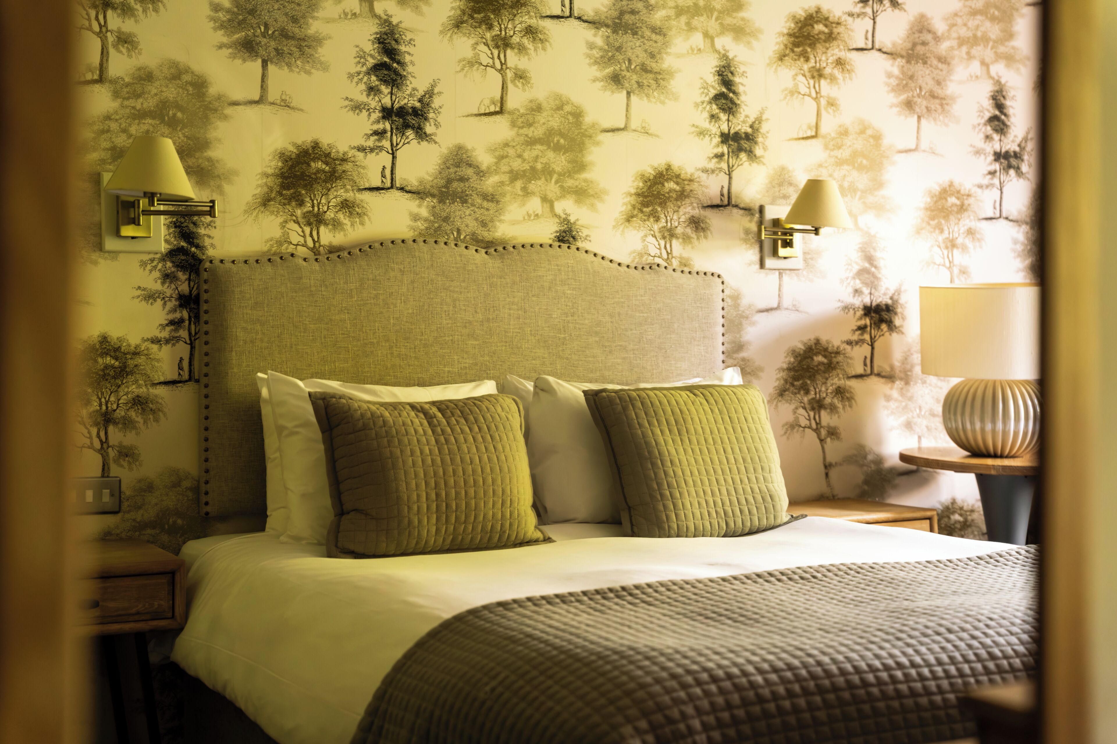 A bedroom with a bed, pillows, a nightstand, a lamp, and a wall with a tree pattern.