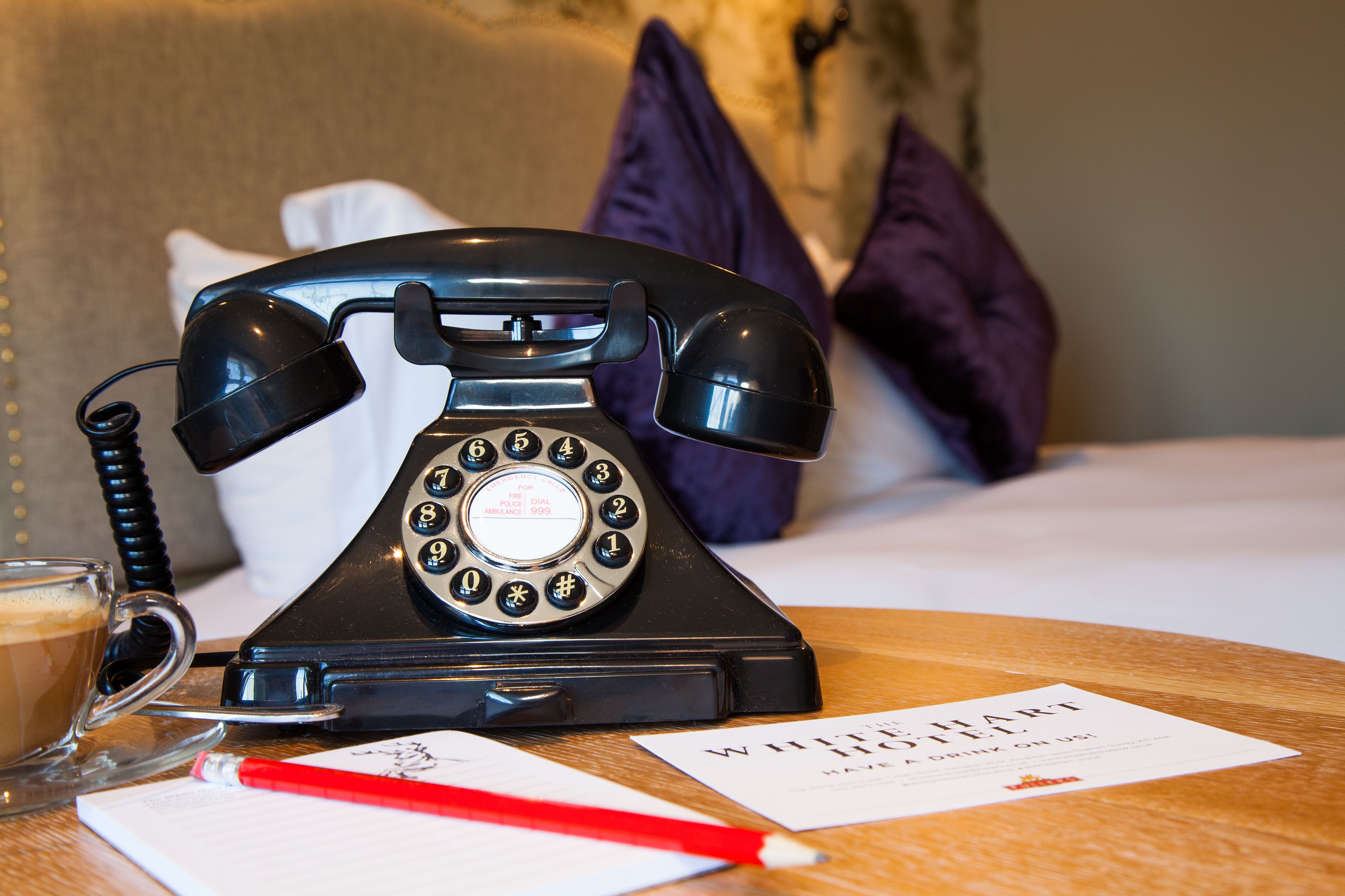 A vintage black rotary dial telephone on a wooden table, a glass of coffee, a red pencil, and a business card.