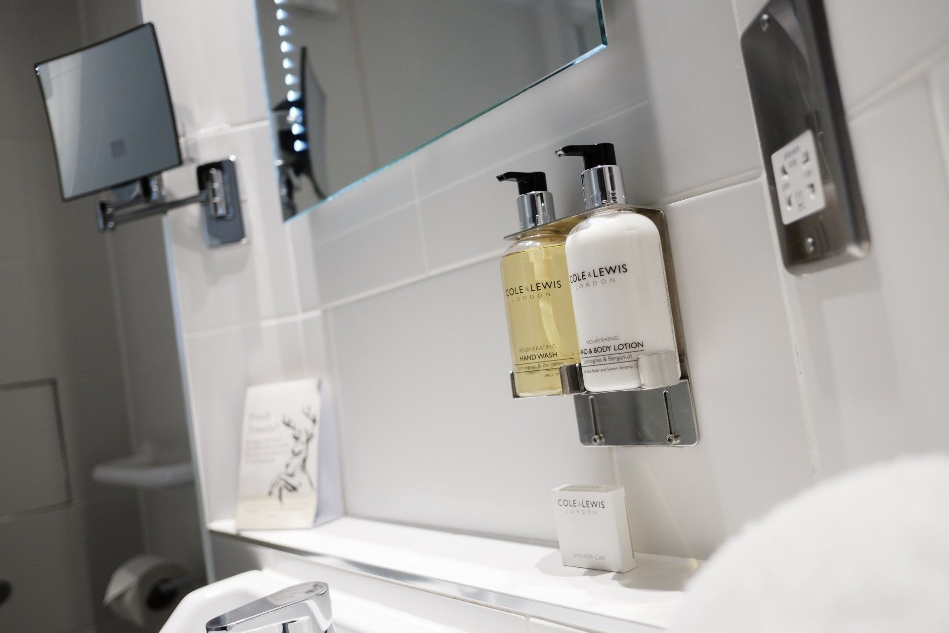 A bathroom with a glass shower enclosure, a mounted TV, and toiletries including hand wash, body lotion, and a shower cap.