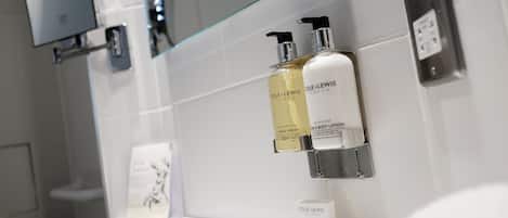 A bathroom with a glass shower enclosure, a mounted TV, and toiletries including hand wash, body lotion, and a shower cap.