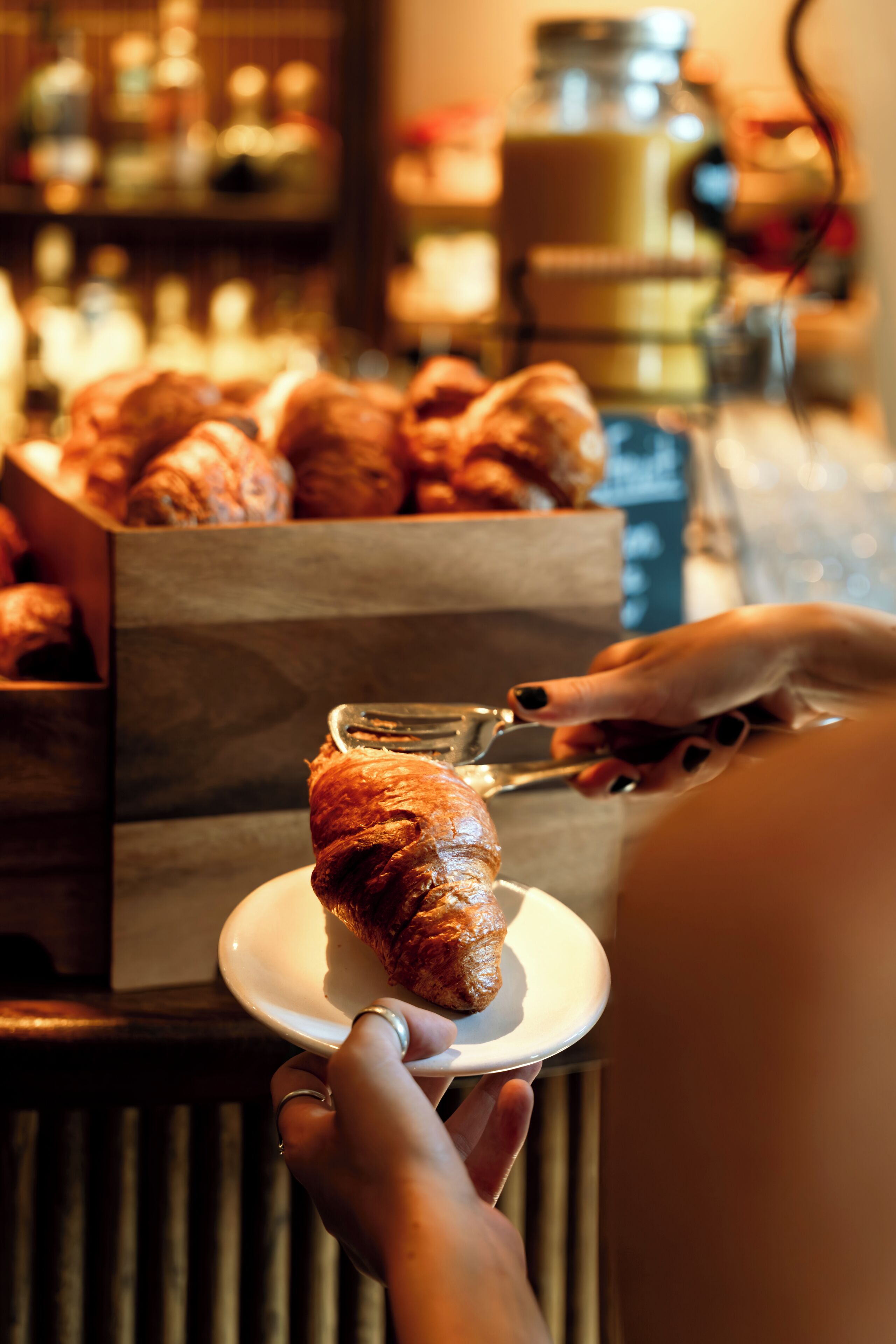 En person håller en croissant på en tallrik, och i bakgrunden syns en låda med croissanter.