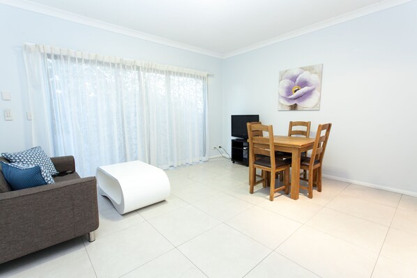 A modern living room with a sofa, a dining table with chairs, a flat-screen TV, and a large window with sheer curtains.