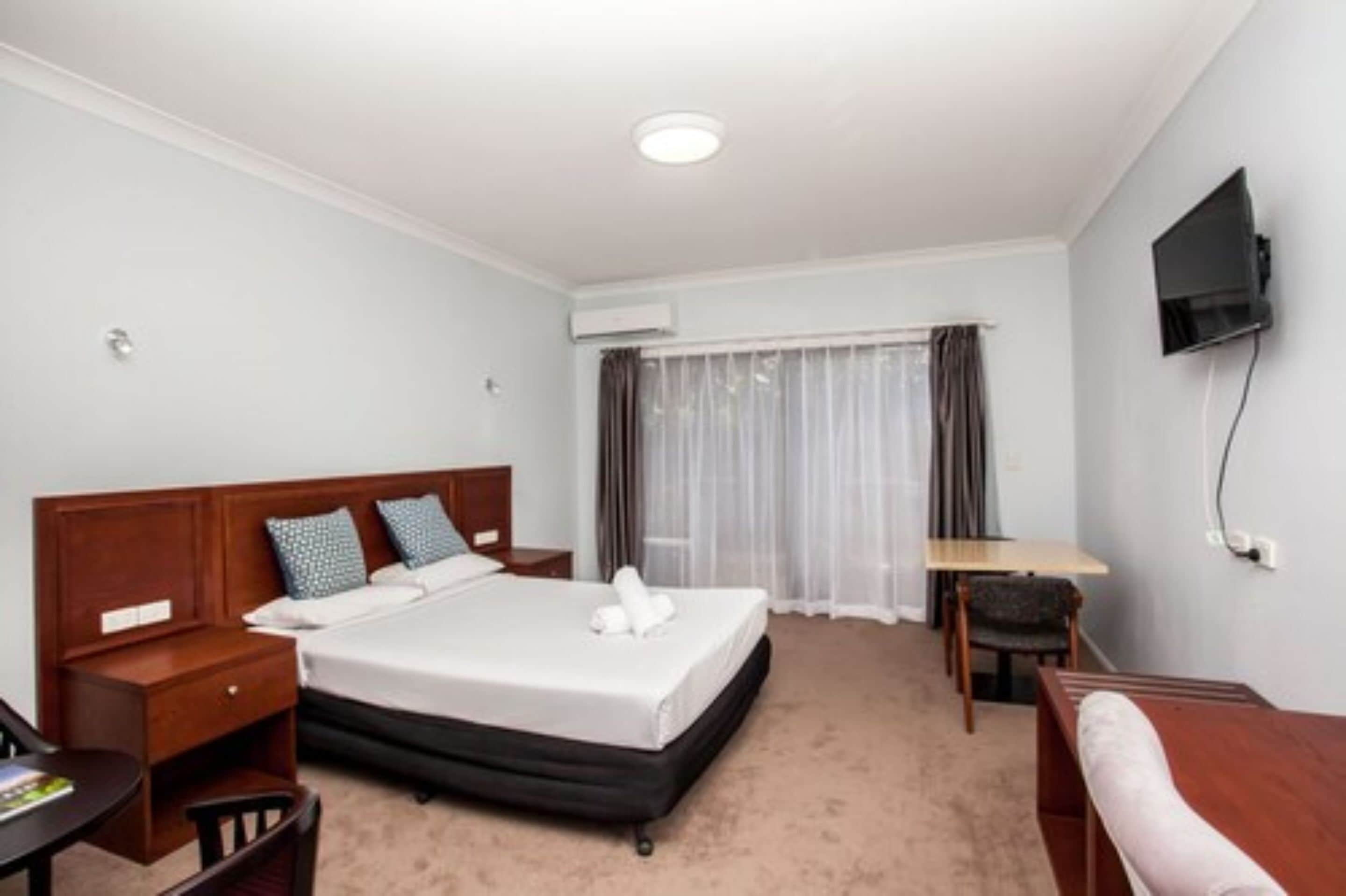 Une chambre d’hôtel comprenant un lit, une table de chevet, une chaise, un bureau, une télévision et une fenêtre avec des rideaux.