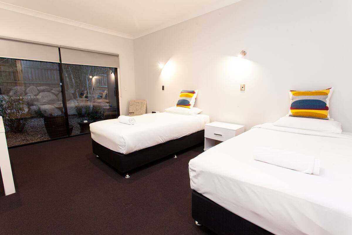 Quarto de hotel com duas camas, uma grande porta de vidro deslizante para um jardim e uma pequena mesa com abajur.