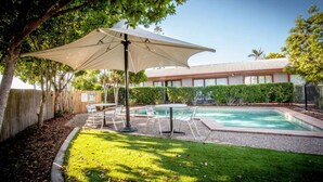 Un patio trasero con piscina, un porche cubierto con sillas y un área cercada con árboles.
