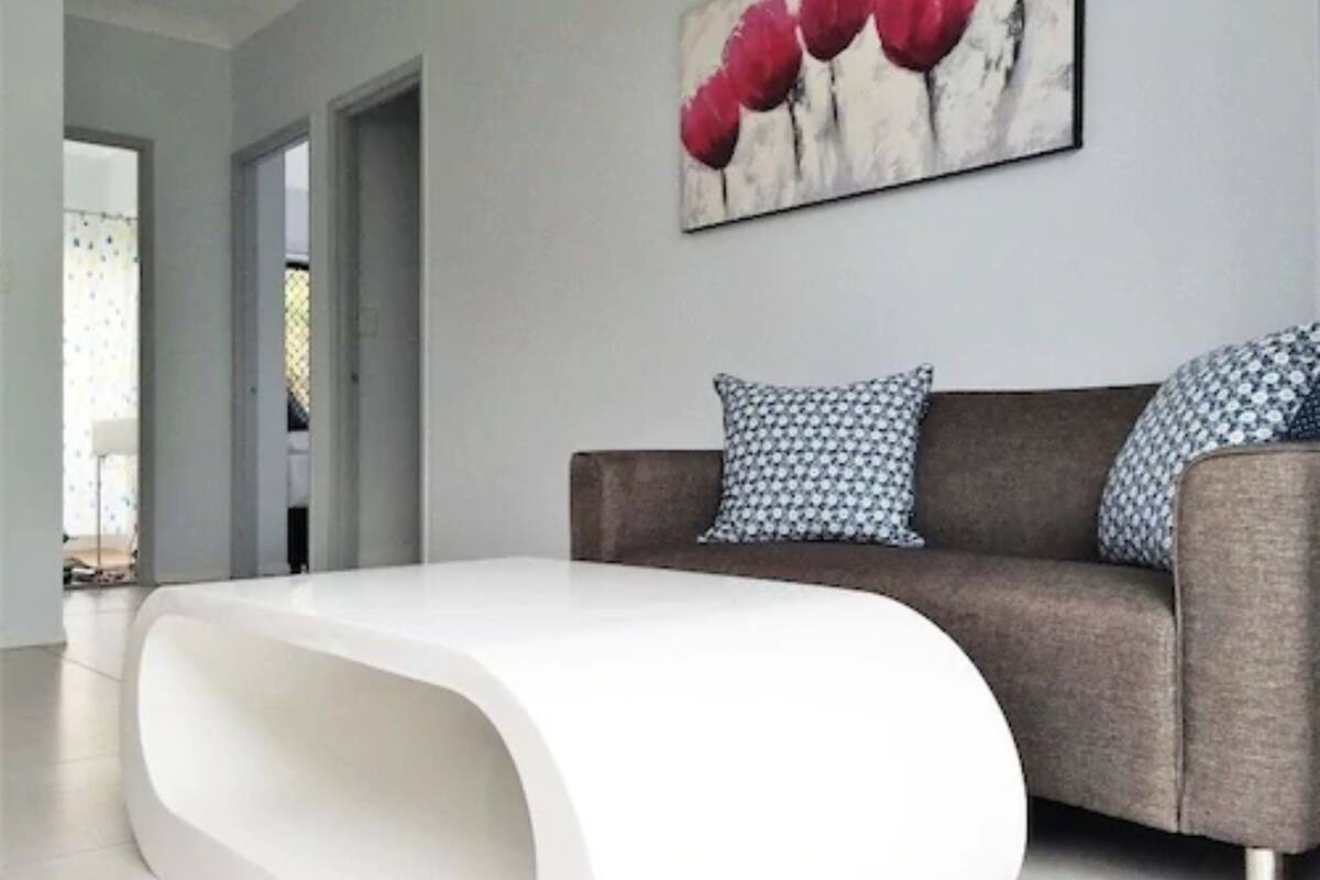 Uma sala de estar moderna com uma mesa de centro branca e curvada, um sofá marrom com almofadas estampadas e um quadro com flores vermelhas na parede.