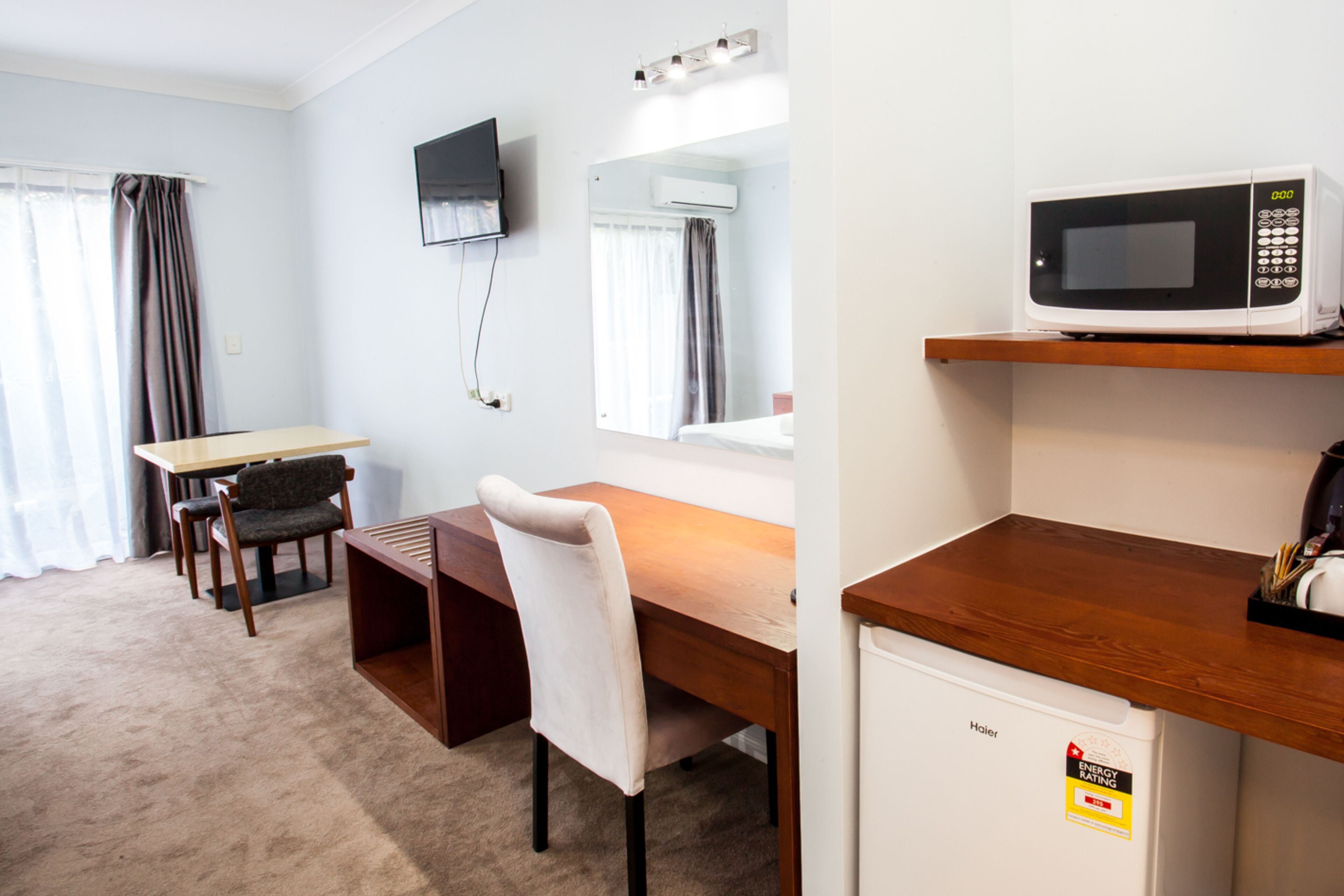 A hotel room with a wooden desk, a white chair, a television mounted on the wall, a microwave on a shelf, and a small refrigerator.