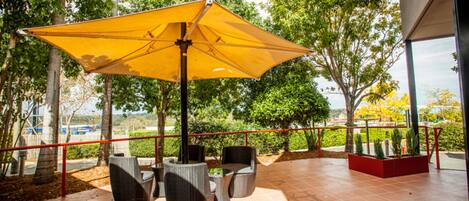 An outdoor patio with wicker furniture and a yellow umbrella, surrounded by trees and a red railing.