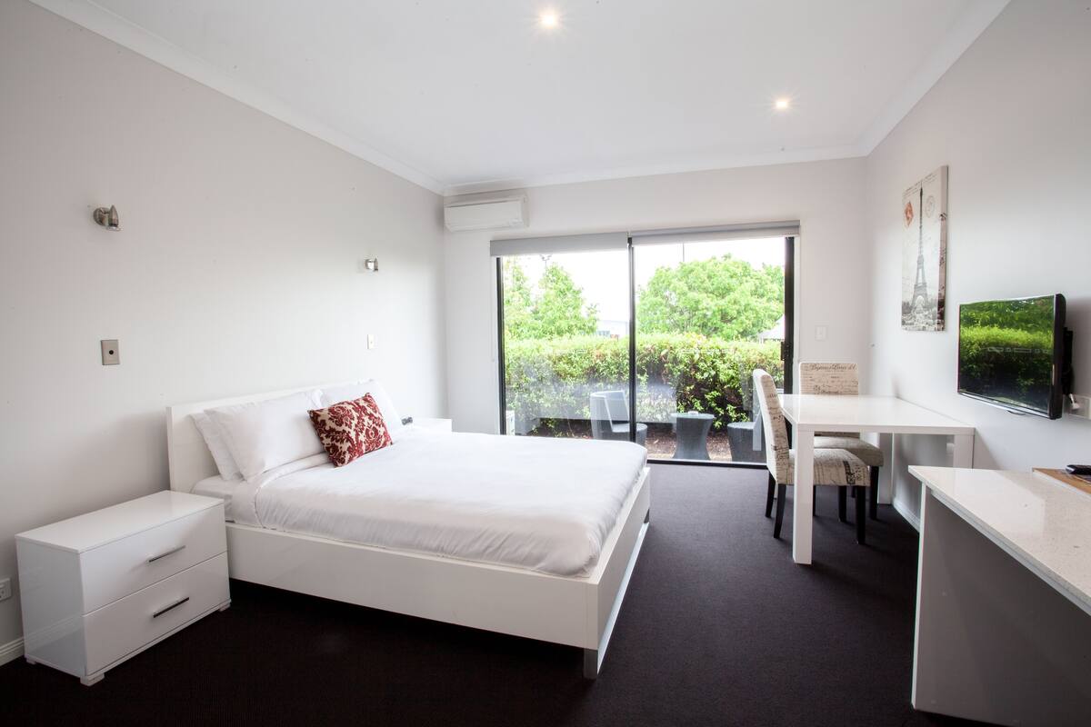 Quarto moderno com uma cama grande, uma escrivaninha e uma cadeira. Há uma janela com vista para o verde lá fora.