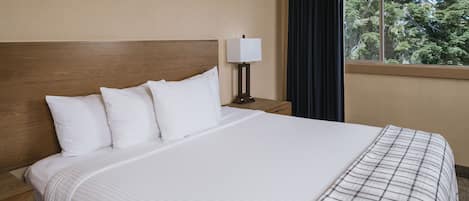 A hotel room with a large bed, a nightstand with a lamp, and a window with a view of trees.
