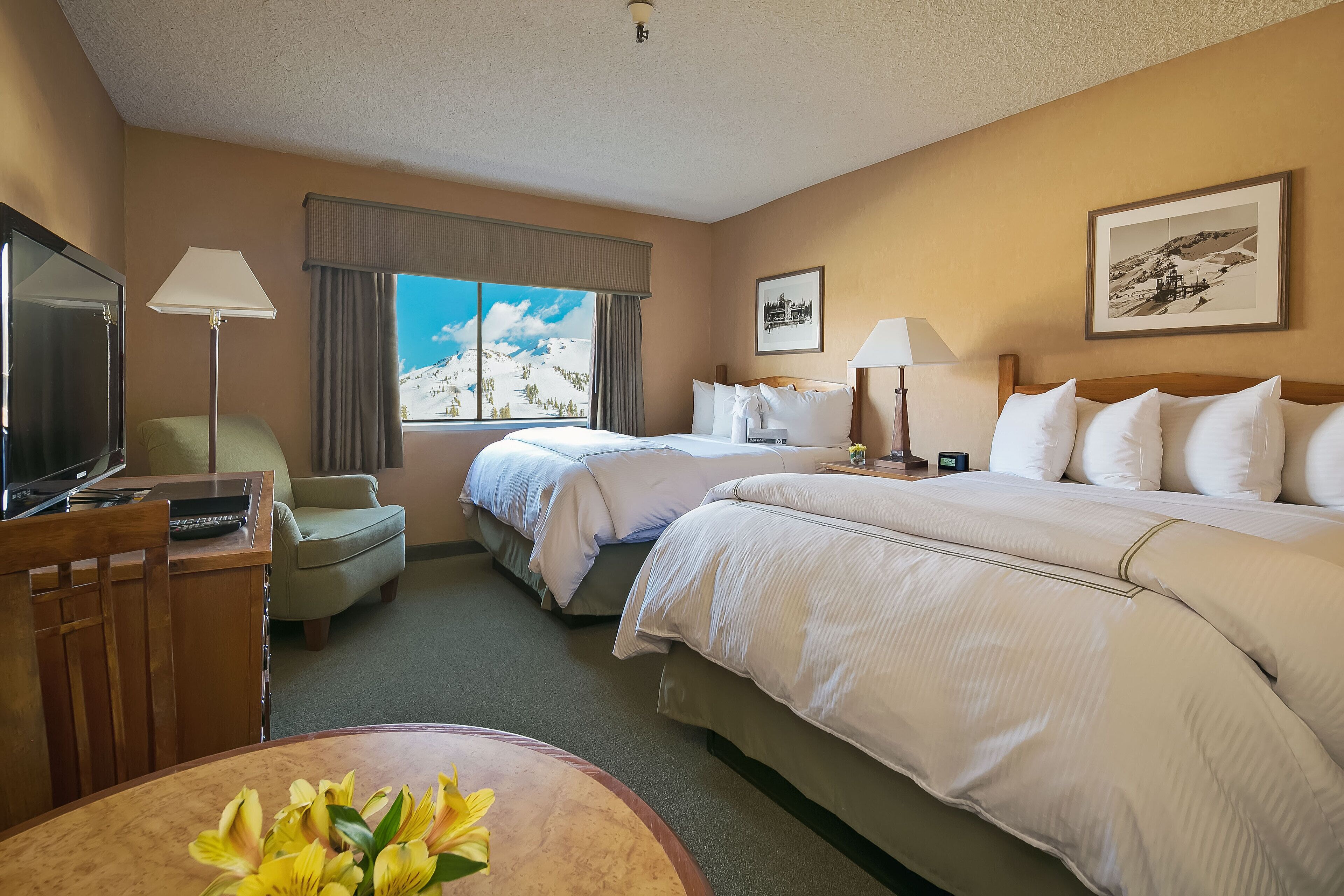 A hotel room with two beds, a desk with a TV, a chair, a window with a mountain view, and a framed picture on the wall.