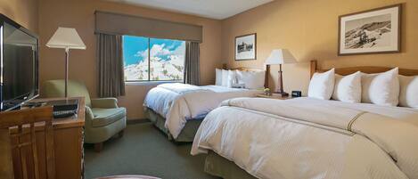 A hotel room with two beds, a desk with a TV, a chair, a window with a mountain view, and a framed picture on the wall.