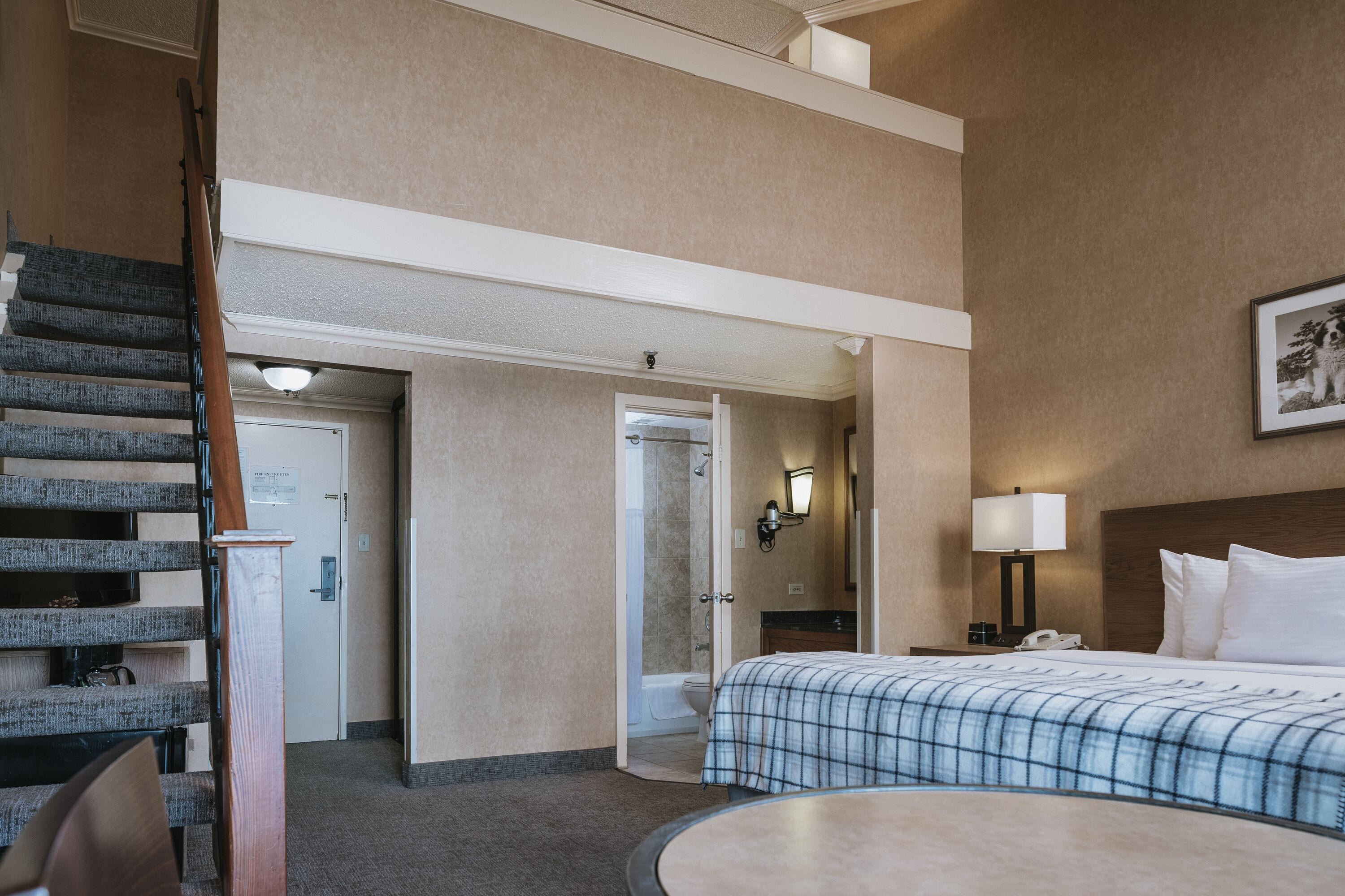 Una habitación de hotel con cama, una mesa redonda, una escalera y un baño visible a través de una puerta abierta.