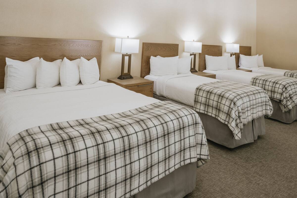 A hotel room with three beds, each with a wooden headboard and white bedding. There are bedside tables with lamps and a carpeted floor.