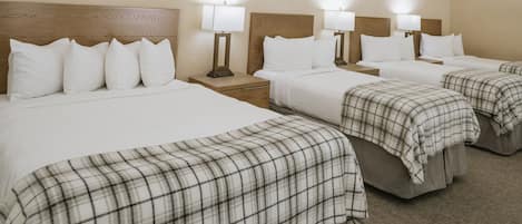 A hotel room with three beds, each with a wooden headboard and white bedding. There are bedside tables with lamps and a carpeted floor.
