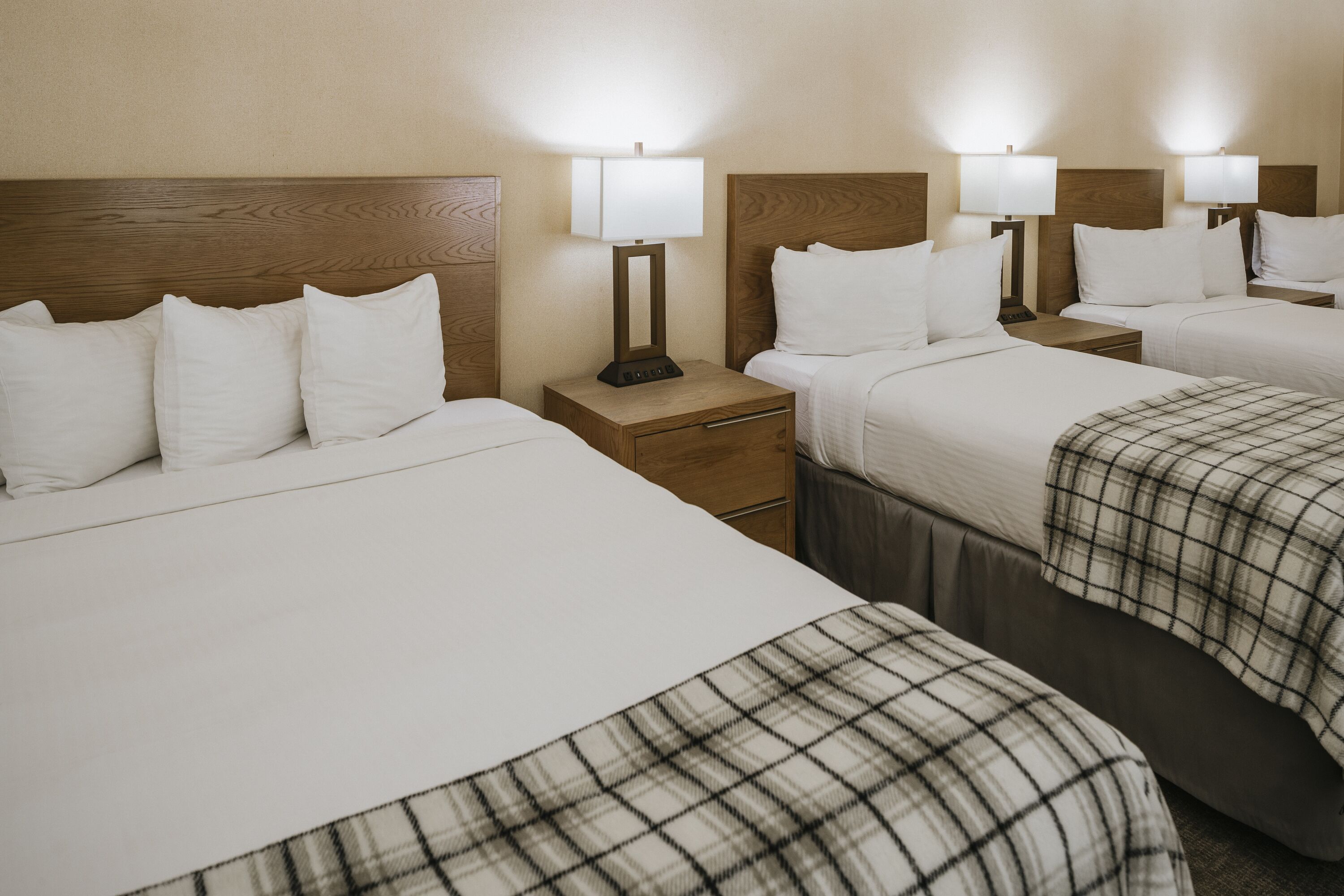 Una camera d'albergo con quattro letti, ognuno con biancheria bianca e una coperta a quadri. Sono presenti comodini con lampade e un poggiatesta in legno.