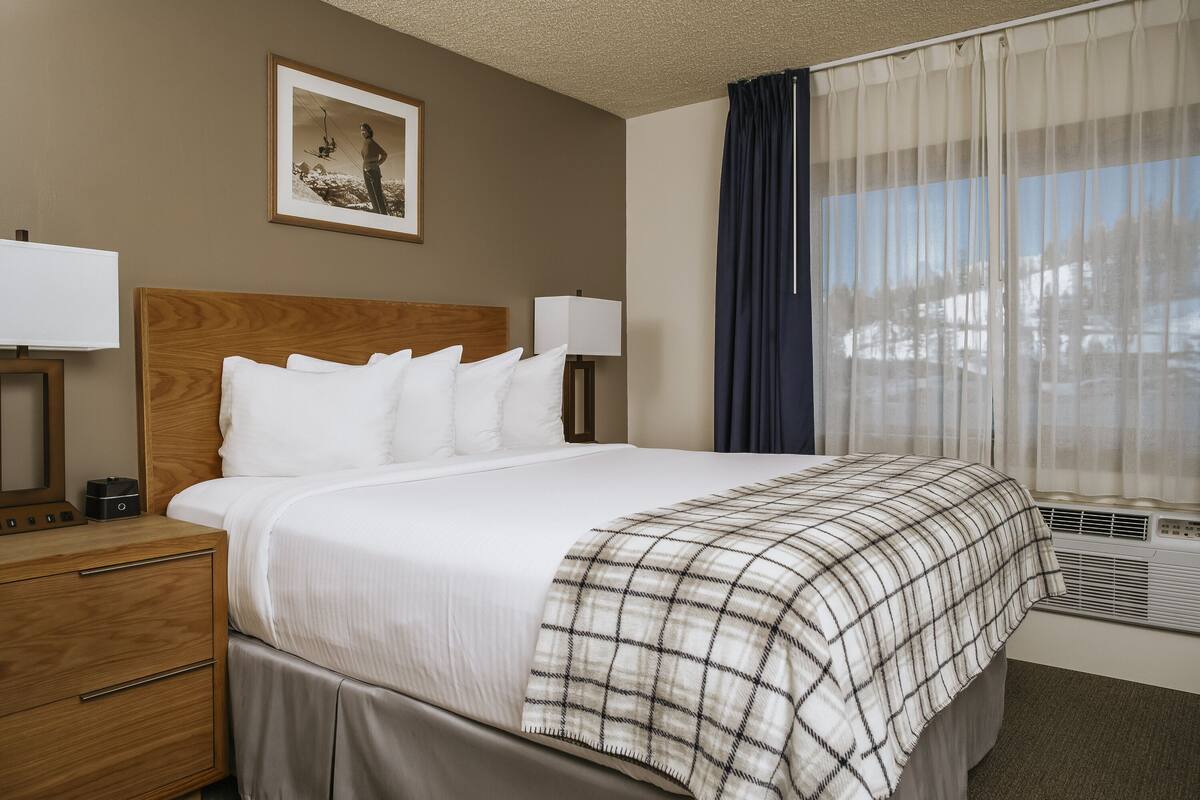 A hotel room with a bed, bedside lamps, a nightstand, a framed picture on the wall, and a window with curtains.