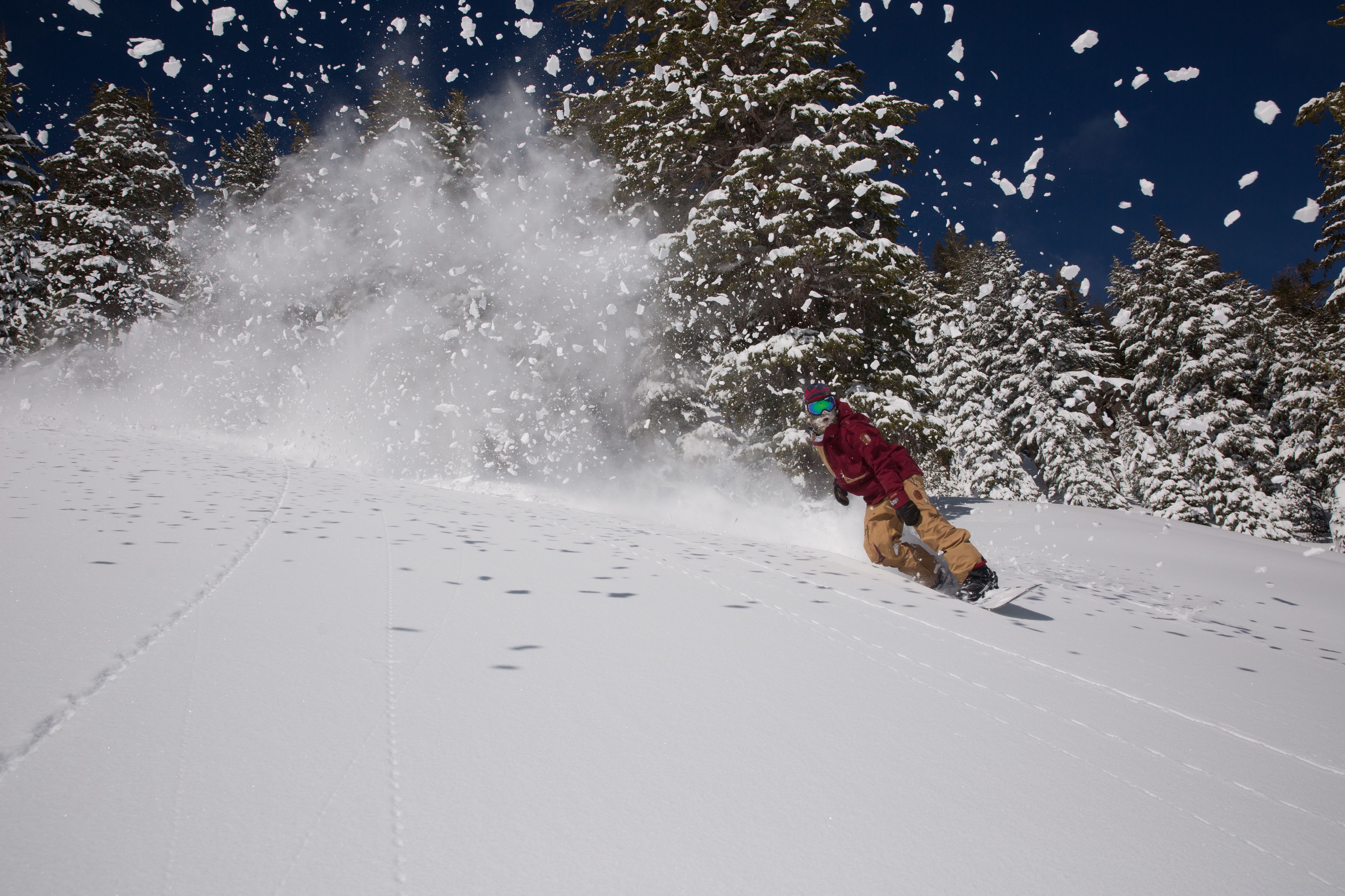 A snowboarder in a red jacket and yellow pants on a snowy slope with trees in the background.
