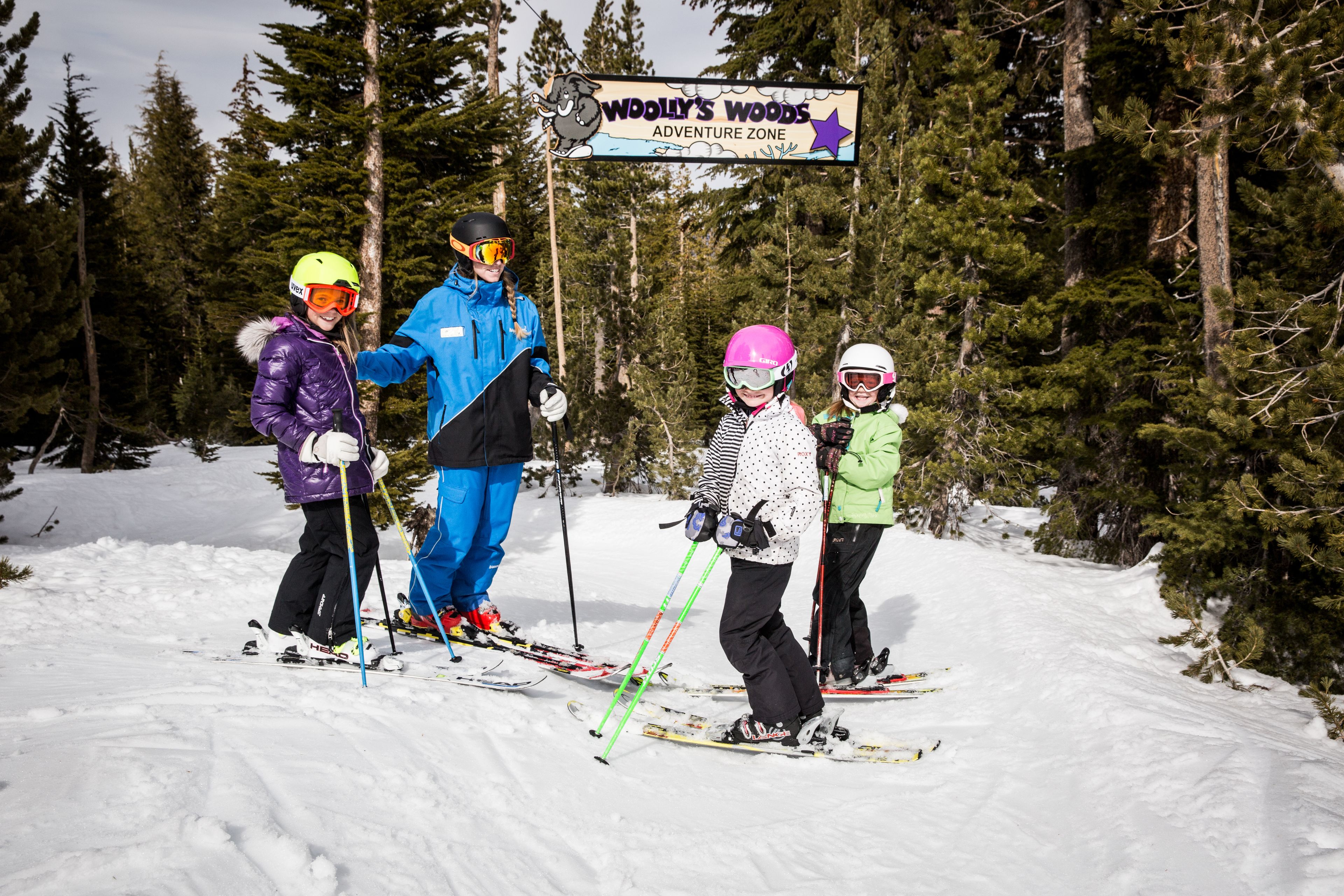 A group of people on skis at a ski resort with a sign reading "Woolly's Woods Adventure Zone".