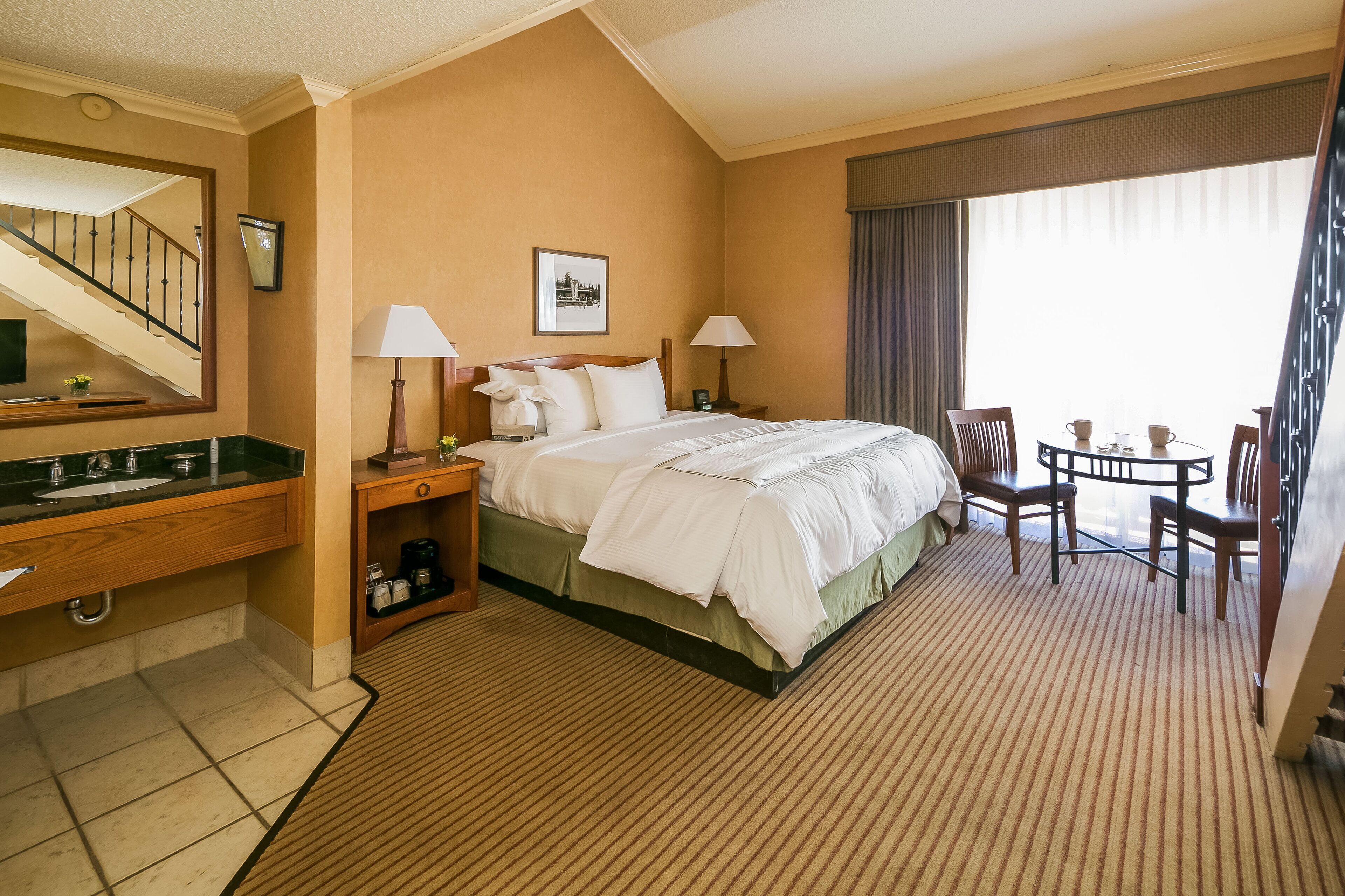 A hotel room with a bed, bedside tables, a desk, a chair, a small table, and a window with curtains.