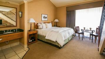 A hotel room with a bed, bedside tables, a desk, a chair, a small table, and a window with curtains.