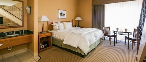 Una habitación de hotel con cama, mesitas de noche, escritorio, silla, una pequeña mesa y ventana con cortinas.