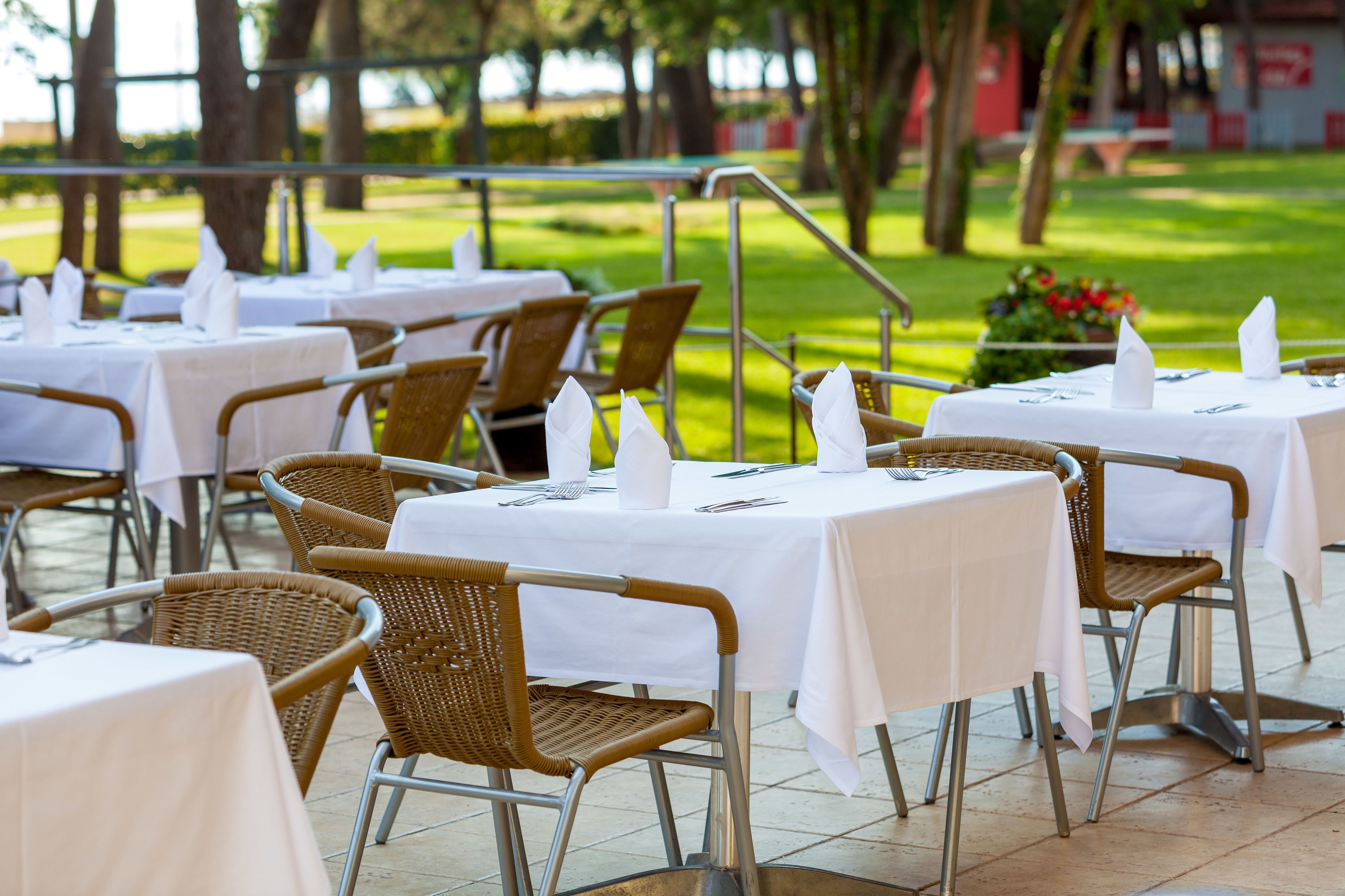 Outdoor dining tables with wicker chairs and white tablecloths, set on a patio with a view of a grassy area and trees.