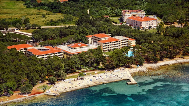 An aerial view of a coastal hotel with multiple buildings, a beach, and a pier. - Hotel Aurora Plava Laguna (Umag)