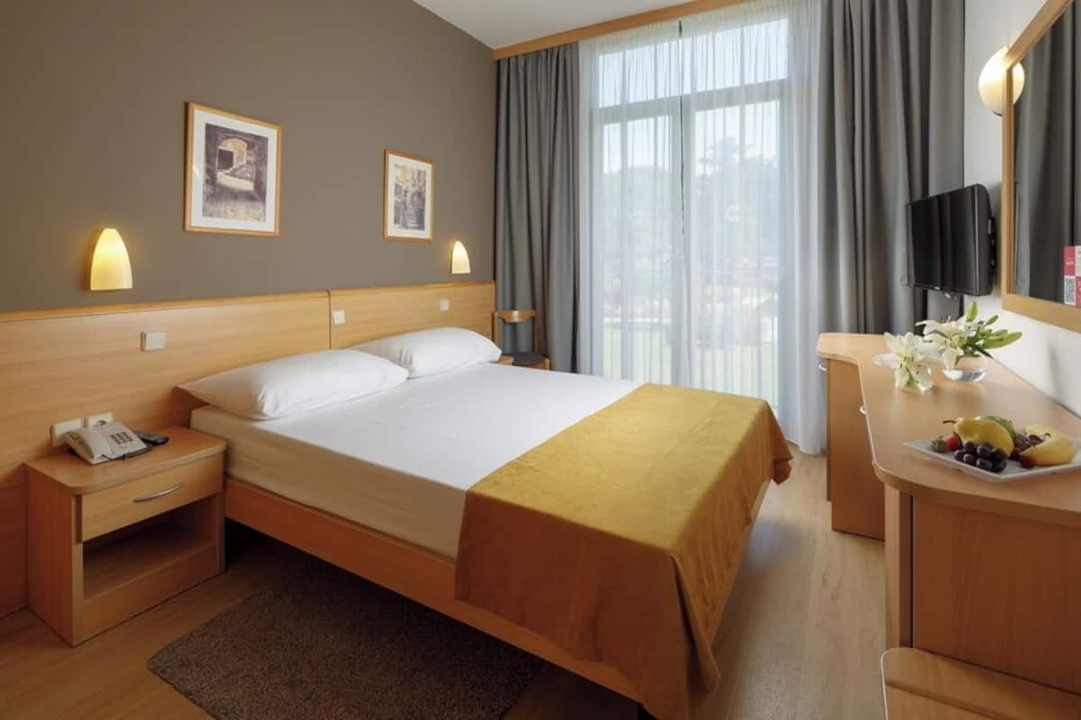 A hotel room with a bed, bedside table with a phone, a desk with a fruit plate, and a TV on the wall.