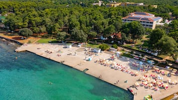 An aerial view of a coastal resort with a beach, a pool, and a large building with solar panels.