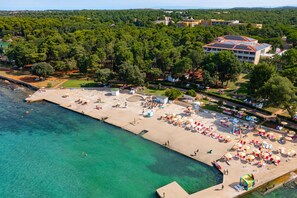 An aerial view of a coastal resort with a beach, a pool, and a large building with solar panels.