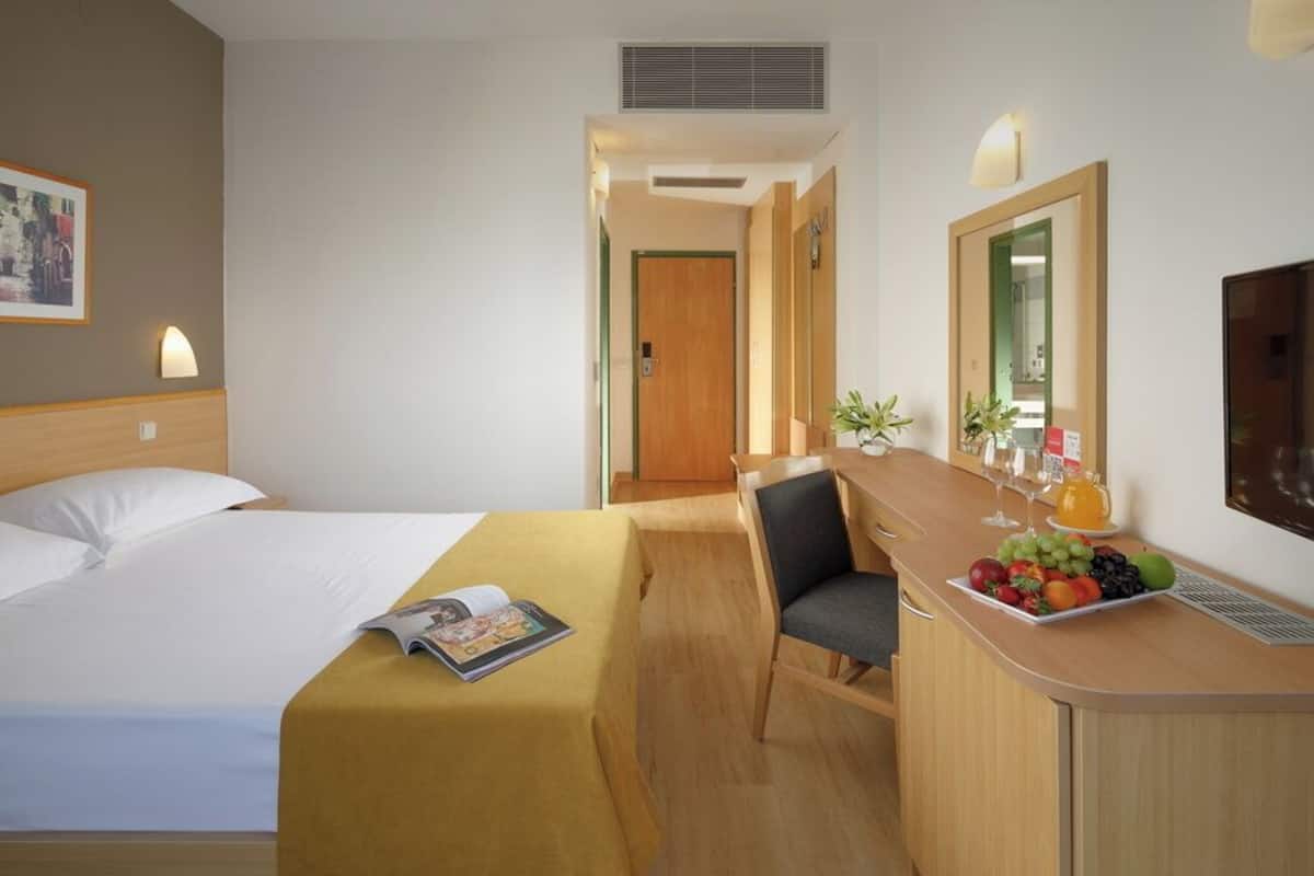 A hotel room with a bed, a desk with a chair, a mirror, and a fruit bowl.