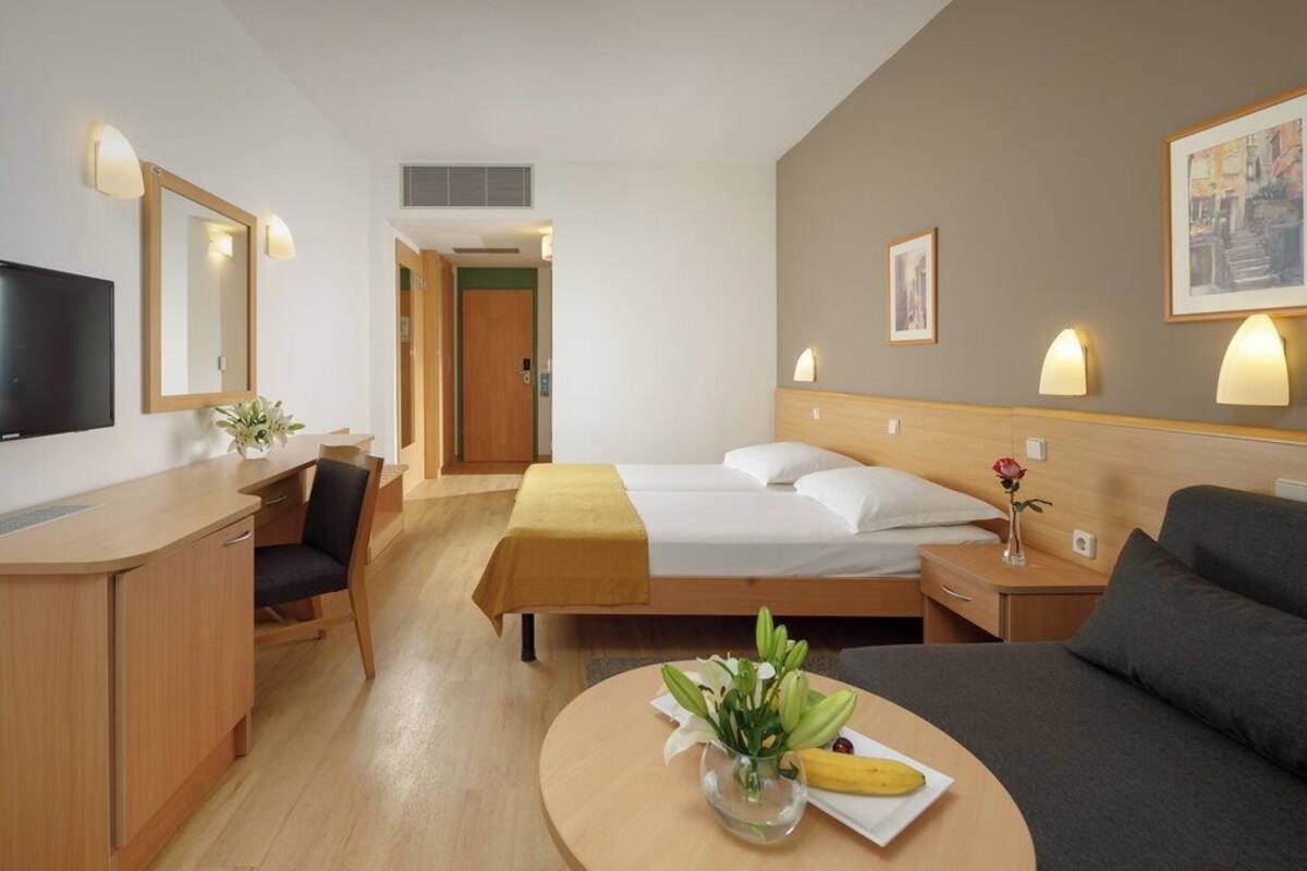 A hotel room with a bed, a desk, a chair, a sofa, a TV, and a small table with a vase and a banana.