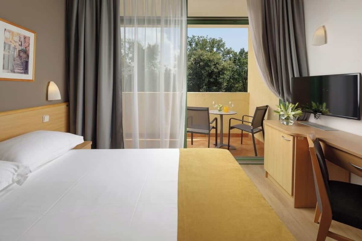 A hotel room with a bed, a desk, a chair, a television, and a balcony with a table and chairs.