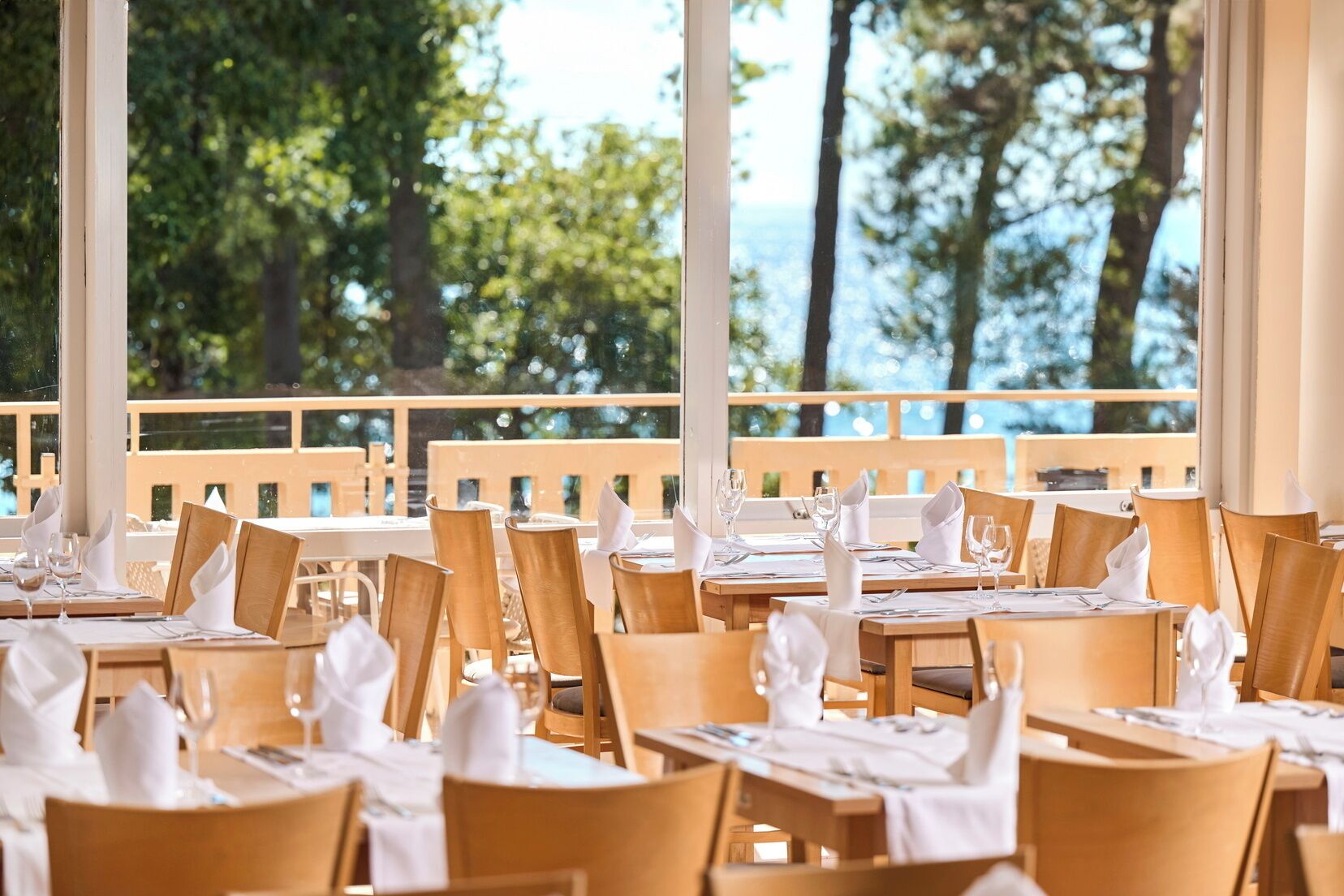 A restaurant with wooden tables and chairs, white tablecloths, and a view of trees and water.