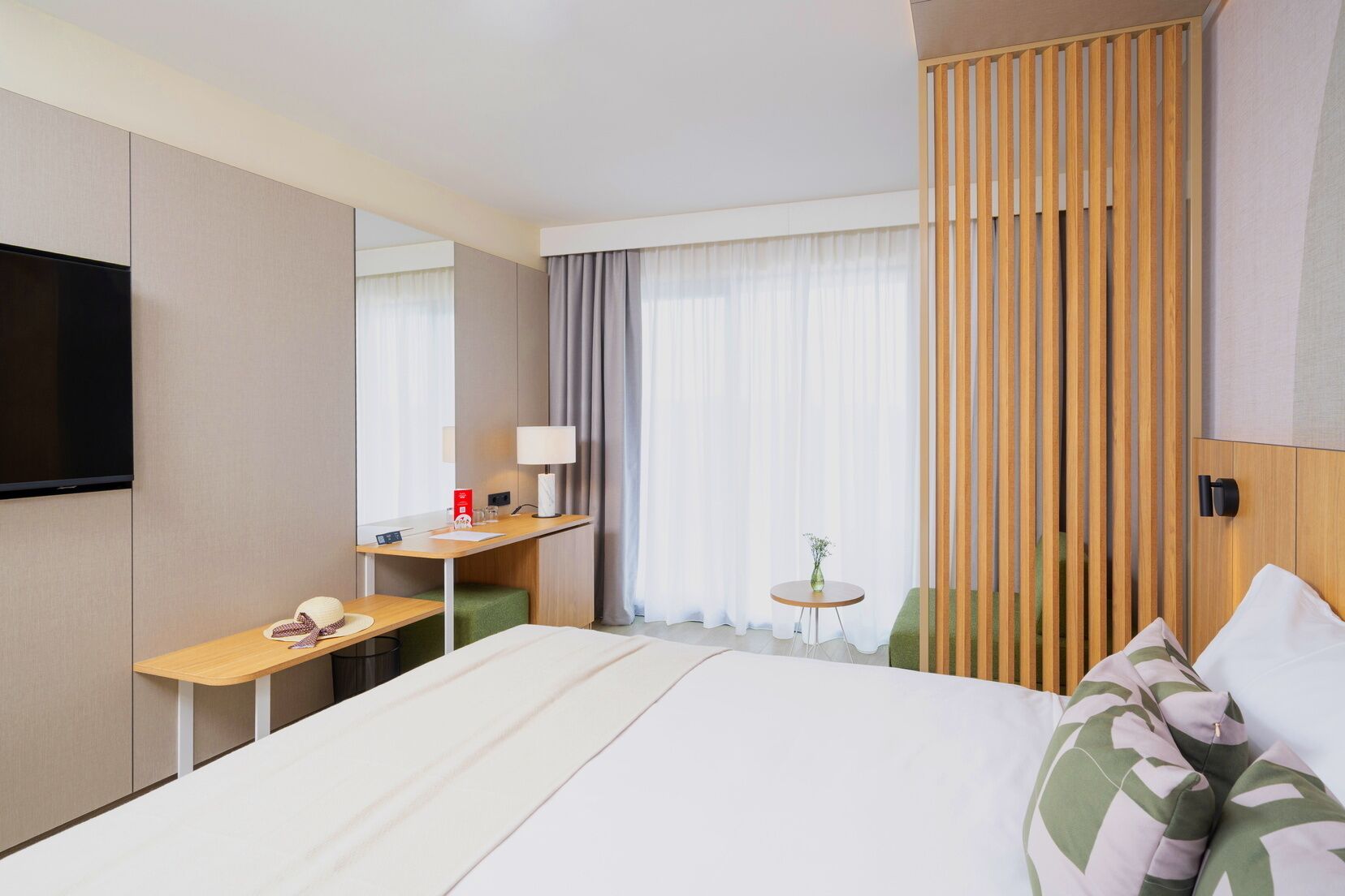 Ein Hotelzimmer mit einem Bett, einem Schreibtisch, einem Fernseher, einem kleinen Tisch mit einer Vase und einem Fenster mit Vorhängen.